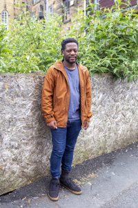 Young Black man standing in front of a wall and some greenery, wearing an orange jacket and blue jeans.